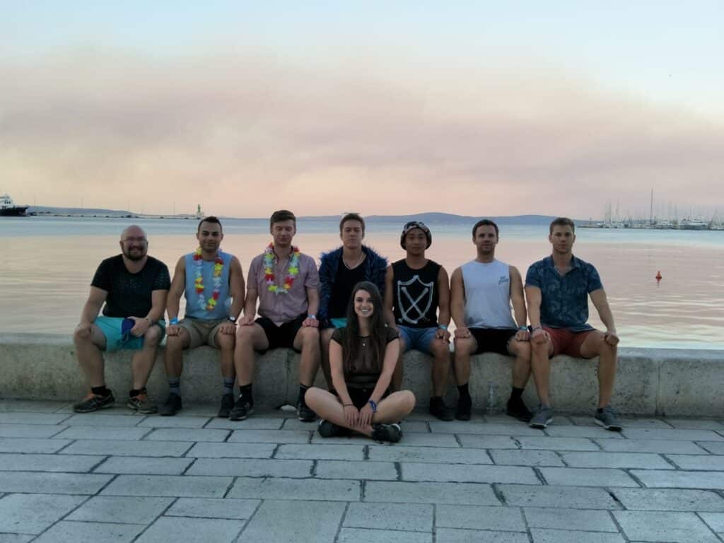 Ultra Music Festival Croatia 5 A group of 8 people sitting on the waters edge posing for a photo. These people have just all attended a music festival for 8 hours.