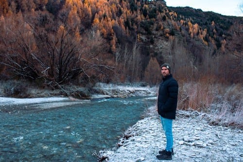A photo of a man standing next to the river in Arrowtown. He has turned to face the camera, the river is next to him and in the background the trees are coloured with orange and red leaves.