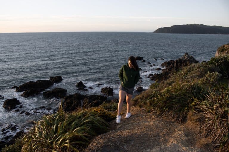 A woman is in the foreground standing near the edge of a cliff at Rocky Bay in Wellington. She is walking towards the camera. The setting sun is off camera but the orange glow is shining on the green plants and gravel path.