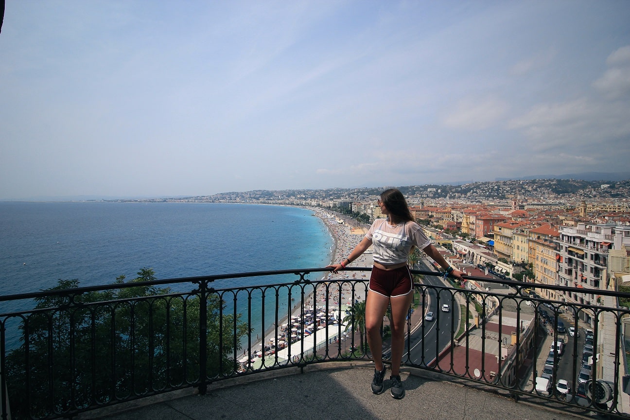 A woman leaning on the bannister at Tour Bellanda in Nice, France. The womans body is facing the camera but her head is turned away to look at the view. The view behind is of Nice beach, the water is bright blue and stretches out to the horizon.