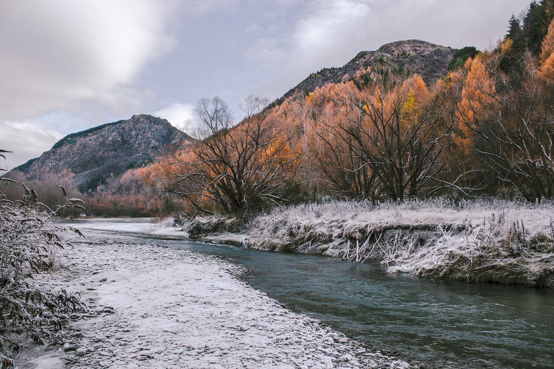 The arrow river in Arrowtown surrounded by snowy, there are a line of trees that have turned orange in the autumn.