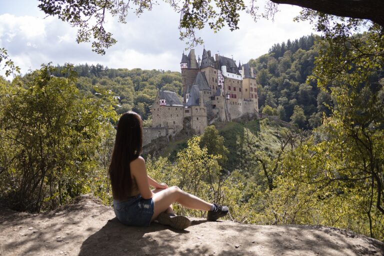 Woman sitting on the edge of a cliff looking over to Burg Eltz Castle in the distance