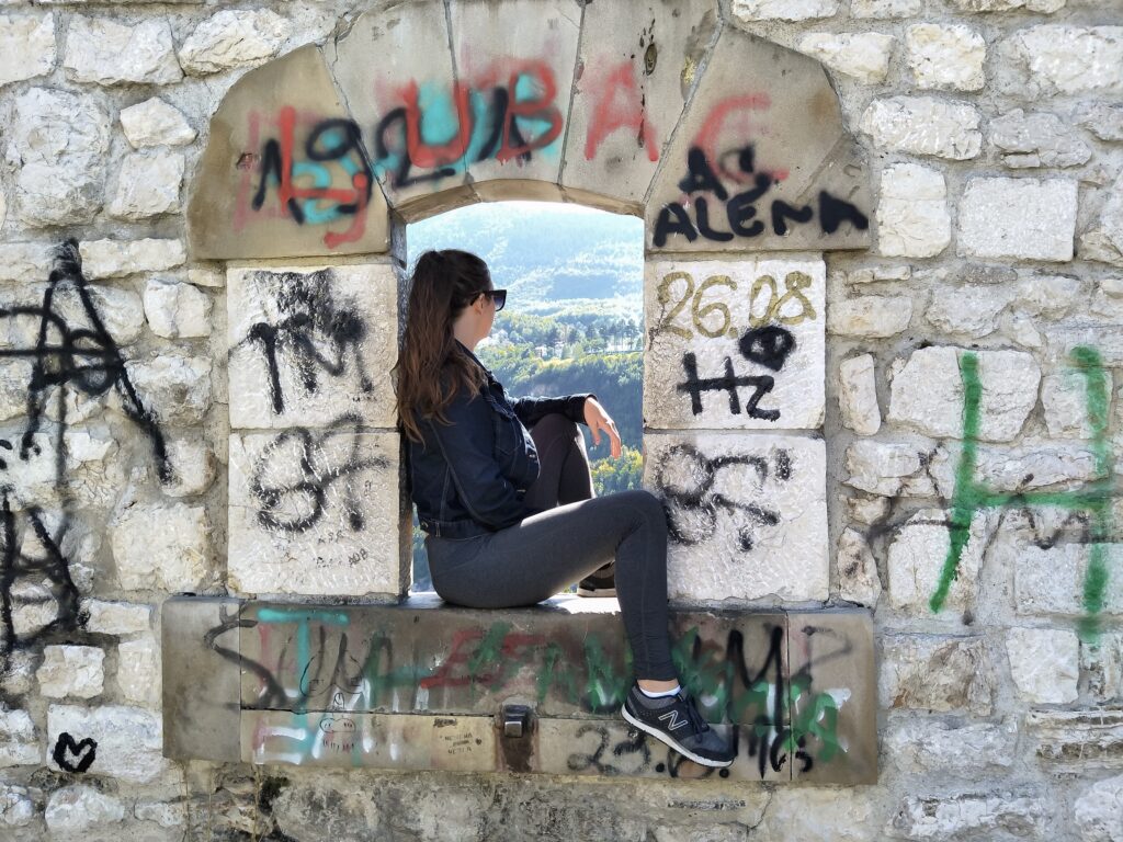 A woman sitting in a window in a block wall looking out to the view. The block wall is covered in spray paint.