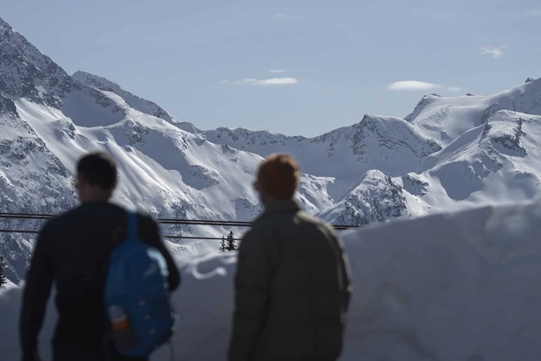 The foreground of the image has a silhouette of two men out of focus, the background is in focus which shows the showy peaks of Whistler mountain in Vancouver.