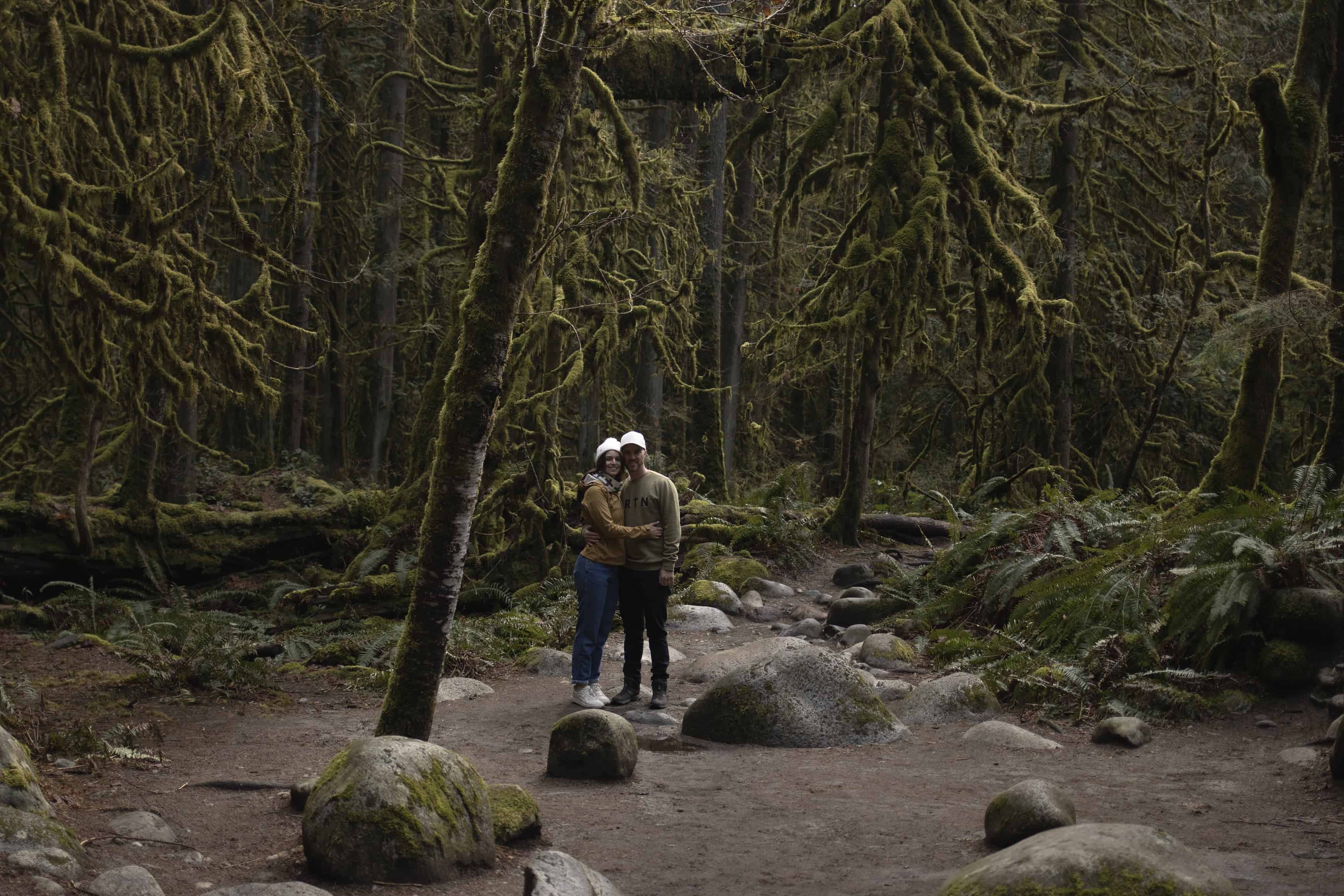 Day trips from Seattle: This photo shows a man and a woman hugging, smiling at the camera while standing among trees at Lynn Canyon Park in Vancouver.