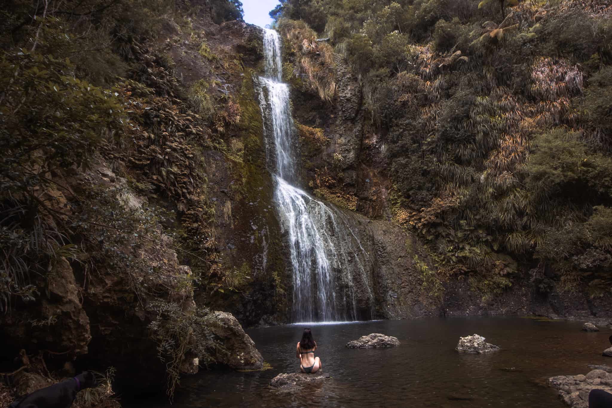 Woman sitting on a rock underneath Kitekite waterfalls in Auckland. The waterfall is 3 tiered and is surrounded by native bush. The rock the woman is sitting on is in the middle of the pool at the bottom of the waterfall. The woman is wearing a bikini and has her back to the camera.