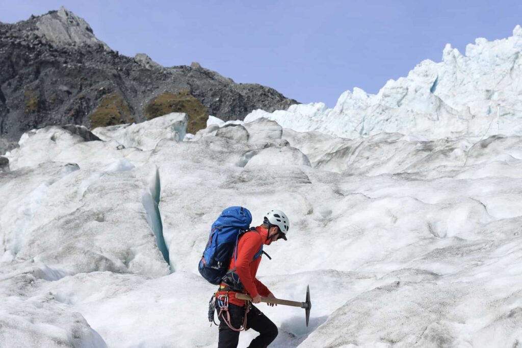 Best Scenic Hikes New Zealand: This image shows the scale of Franz Josef glacier in New Zealand. The foreground has a male hiking up the ice with a pick axe and the background is almost completely the glacier.