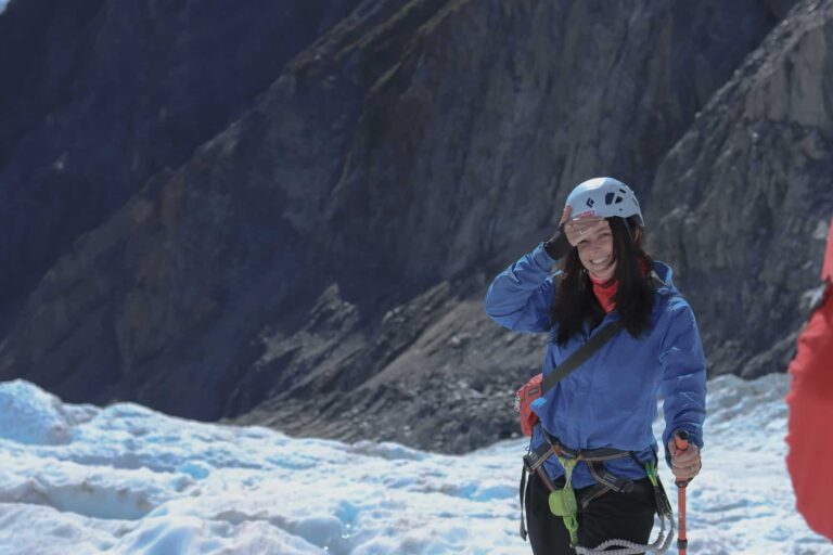 A female smiling at the camera, she is wearing a safety helmet, waterproof jacket and hiking gear. The background shows an icy shelf she is standing on which is a glacier which then slowly falls into the valley below