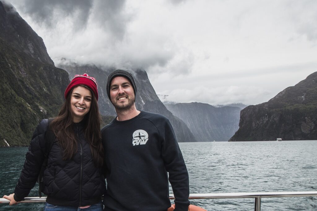 A male and female, standing on the edge of a boat smiling at the camera. In the background is Milford Sound with low lying clouds and large mountains.