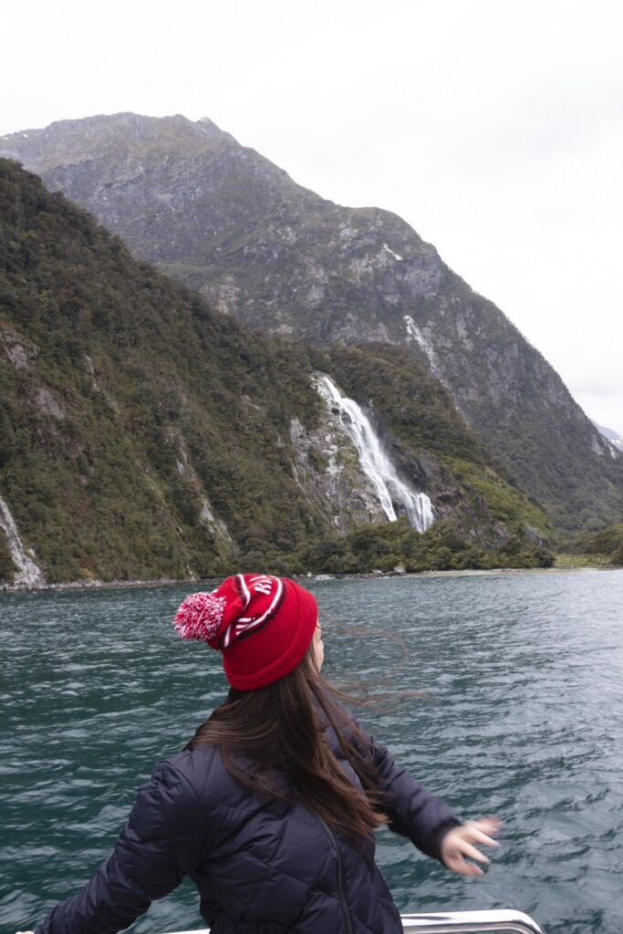 Te Anau to Milford Sound: Milford Sound Cruise 5 Woman standing on a boat, turning around to look at a large waterfall in Milford Sound in the background