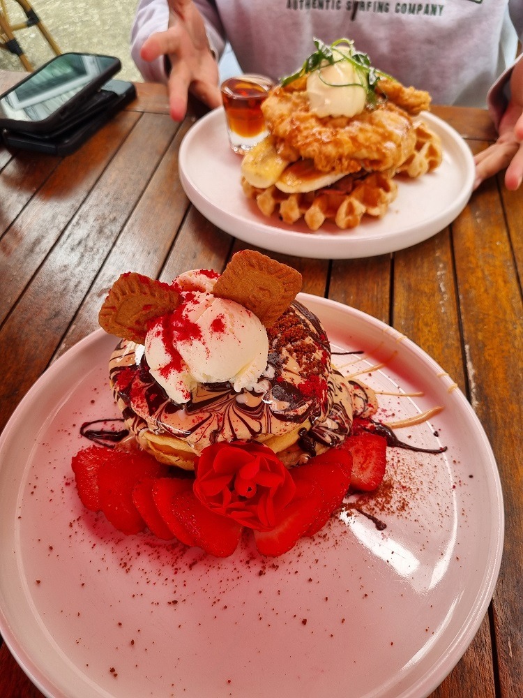 7 of The Best Indian Restaurants in Auckland 11 Image of two plates, one plate is stacked with 2 large fluffy pnackes, pink candy floss, a dollop of ice cream, drizzle of chocolate and caramel sauce and topped with 2 Biscoff cookies. The second plate has a stack of waffles with ice cream, a banana split in half and drizzled with chocolate and caramel sauce.