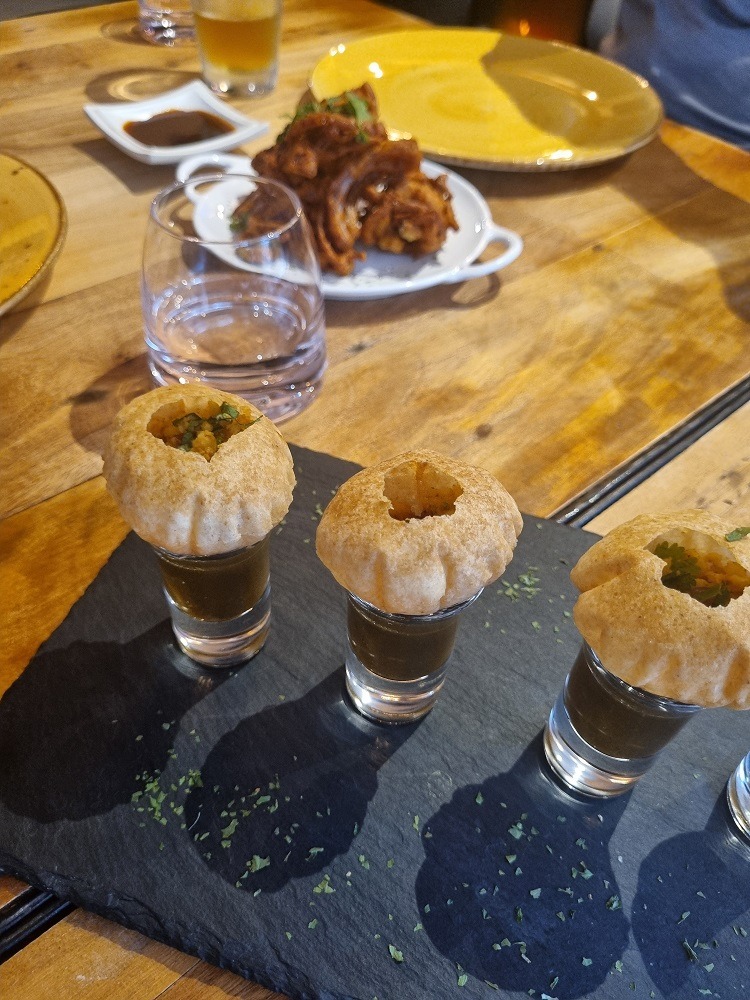 7 of The Best Indian Restaurants in Auckland 7 3 pani puri morsels are balanced on top of a shot glass which are filled with an Indian sauce, in the background is a plate of onion bhaji.