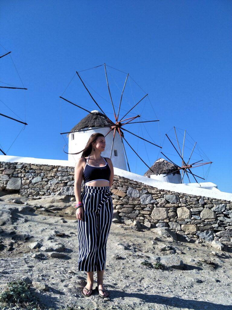 A woman standing in front of the windmills of Mykonos Island, 2 white windmills can be seen behind the woman and 1 can be partially seen in the left edge of the photo.