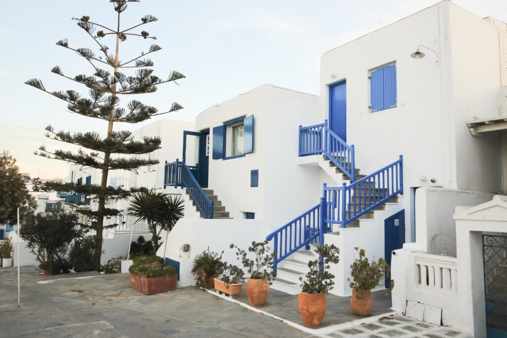The white and blue buildings of Mykonos, Greece. There are pot plants outside the homes with staircases coming down front the front door.