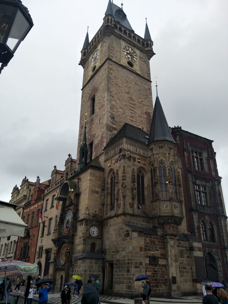 The Prague astronomical clock is in the right side of the photo, there is a large crowd of people standing below the clock.