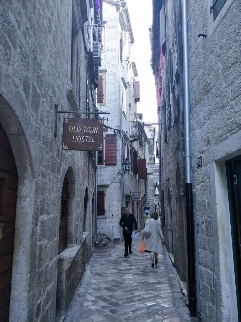 A view where to stay in Kotor Old Town showing the sign for Old Town Hostel. The street is small, and the tall building are built from grey blocks.