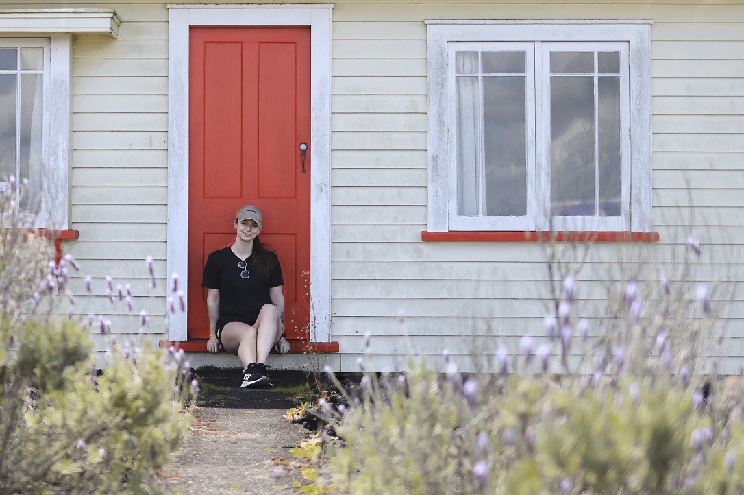A woman sitting in the doorway of a cottage as part of Accommodation on Rangitoto Island. In front of her is a bunch of lavender bushes in full bloom