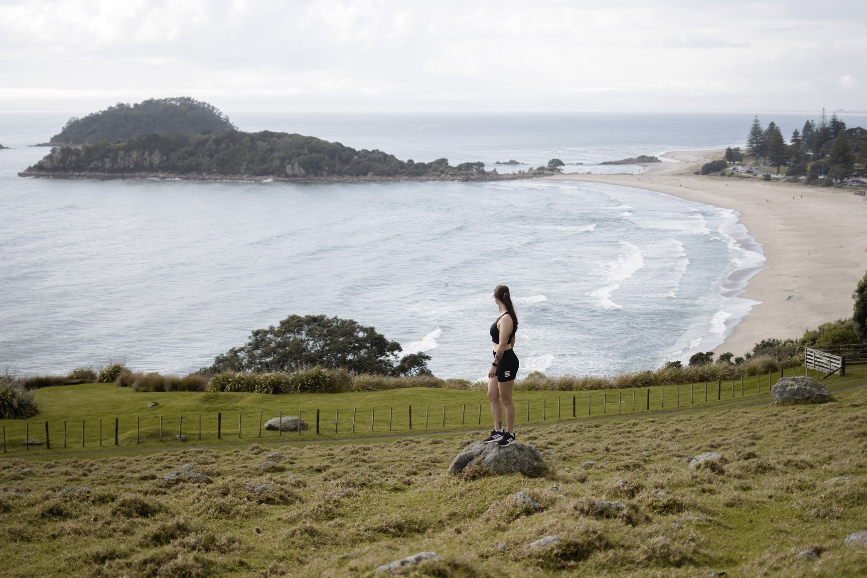 Woman standing on a large rock with Mount Maunganui beach behind her. She is standing near the beginning of one of the Mt Maunganui hiking trails