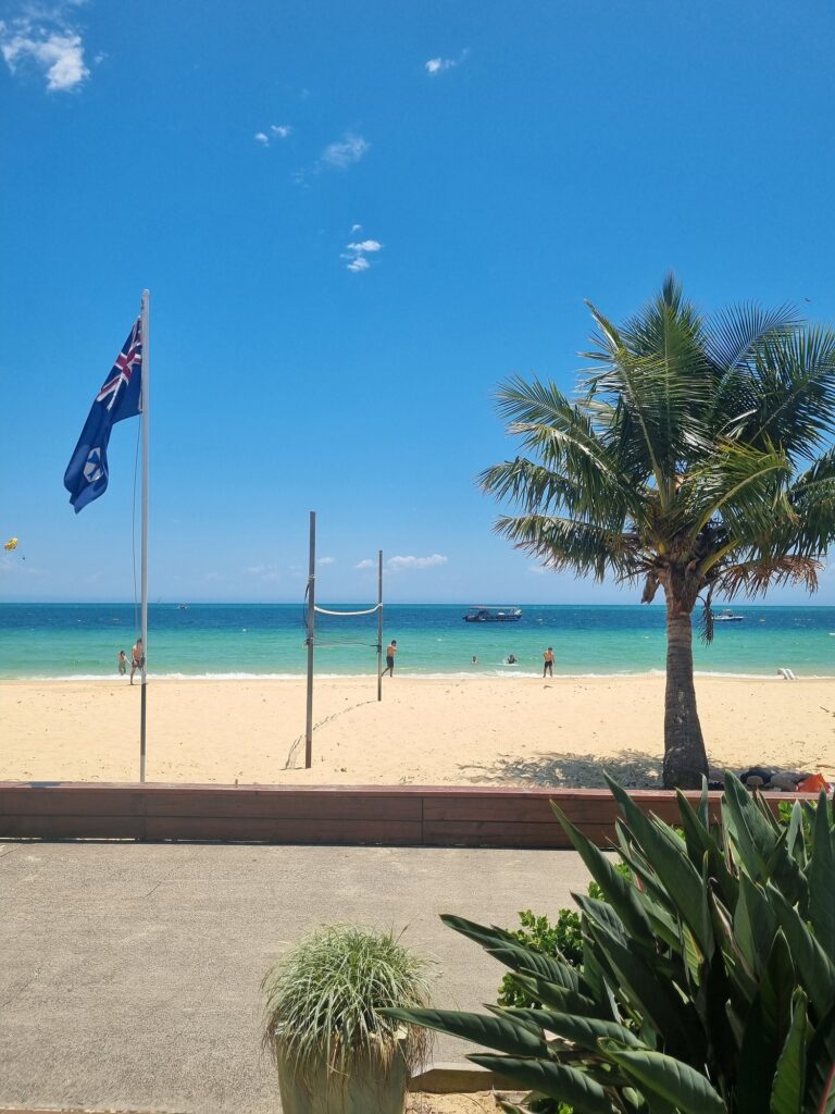 Tangalooma Resort on Moreton Island. This photo shows the white sand beach from the restaurant. A volleyball net is set up and there are people swimming in the clear blue ocean.