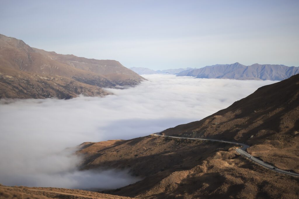 Amazing Queenstown Travel Guide 2026 Views from the top of the Crown Range in Queenstown. Thick fog is covering the lower half of the valley, sun is pouring across the grassy paddocks