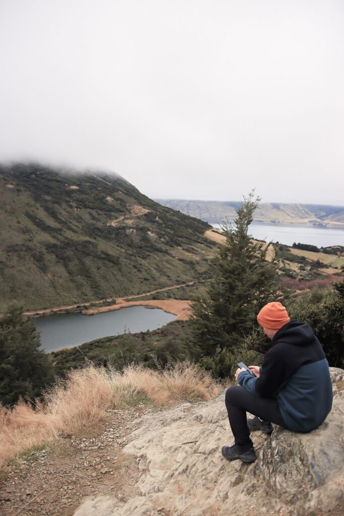 New Zealands Best Scenic Hikes: South Island Male sitting near the edge of a cliff on the Mt Crichton loop track in Queenstown. The Cliff is overlooking Moke Lake below.