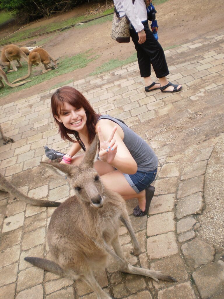 A teenager is crouching on the ground behind a Kangaroo, she is smiling at the camera and pulling the 'peace' sign with her hand. This photo was taken at Currumbin Wildlife Park