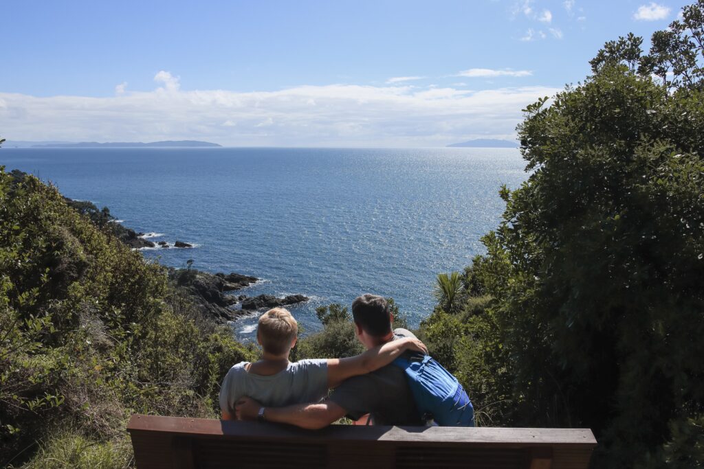 Best Things to do in Auckland 2026 A male and female sitting on a wooden bench overlooking the Hauraki Gulf from Tiritiri Matangi Island