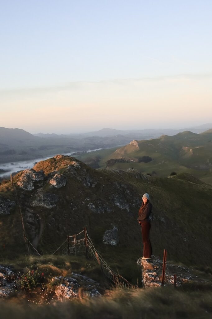 Ultimate 10 Day North Island Itinerary A woman is standing on a rock near the edge of a cliff at Te Mata Peak in Hastings. The orange glow of the sunrise is shining on her face and she is smiling at the camera. The background is of rolling green hills with a light blue and orange sky behind her.