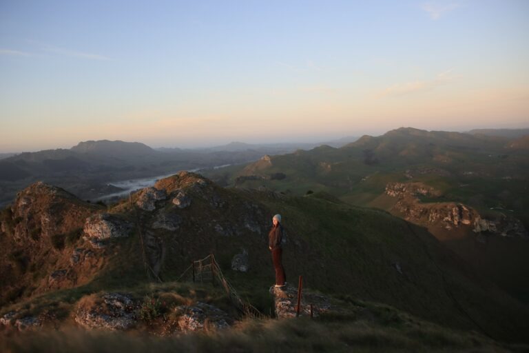 woman is standing on a rock near the edge of a cliff at Te Mata Peak in Hastings. The orange glow of the sunrise is shining on her face and she is smiling at the camera. The background is of rolling green hills with a light blue and orange sky behind her.