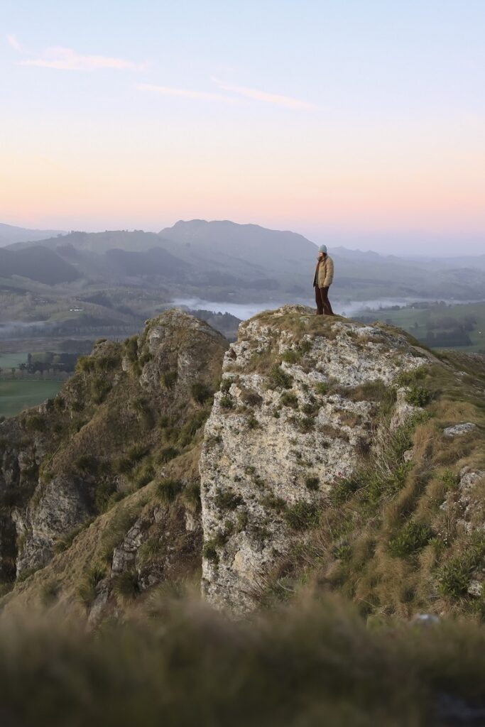 Ultimate 10 Day North Island Itinerary A woman is standing on the edge of the cliff at Te Mata Peak near Napier. The sunrise is glowing behind her and the sky is covered in pink and blue hues. The Cliff she is standing on is a mixture of rock and grassy bush